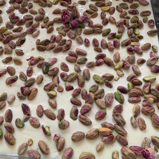 Photo of a white creamy looking desert  covered in pink and green pistachios and a scattering of pink rose petals, a little flower design has also been made from a few small rose buds